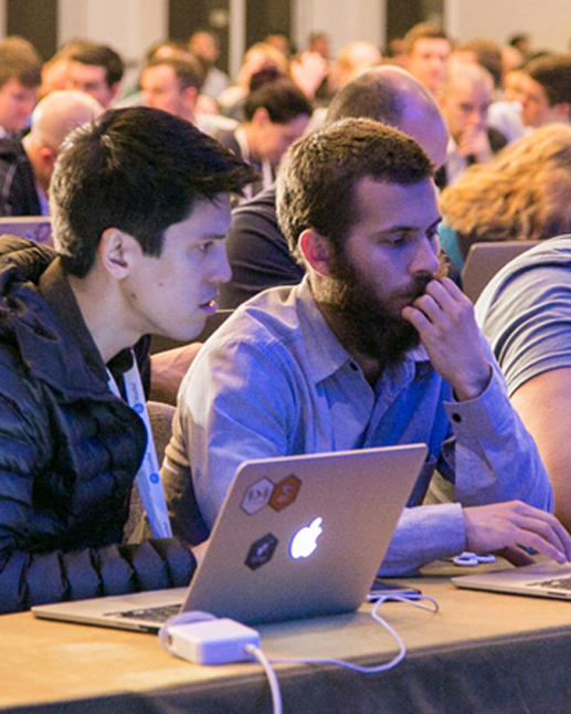 2 men review an offscreen laptop with others sitting at tables in the background at a workshop event