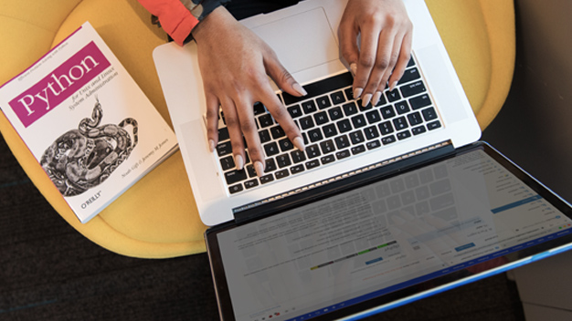 Overhead image of a person typing on their laptop with a Python book to their right